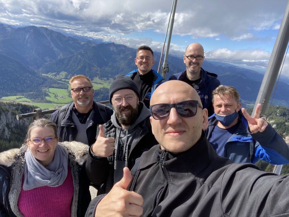 Gruppenselfie des SimplyTest-Teams auf einem Berggipfel bei Bayrischzell, im Hintergrund die Alpen