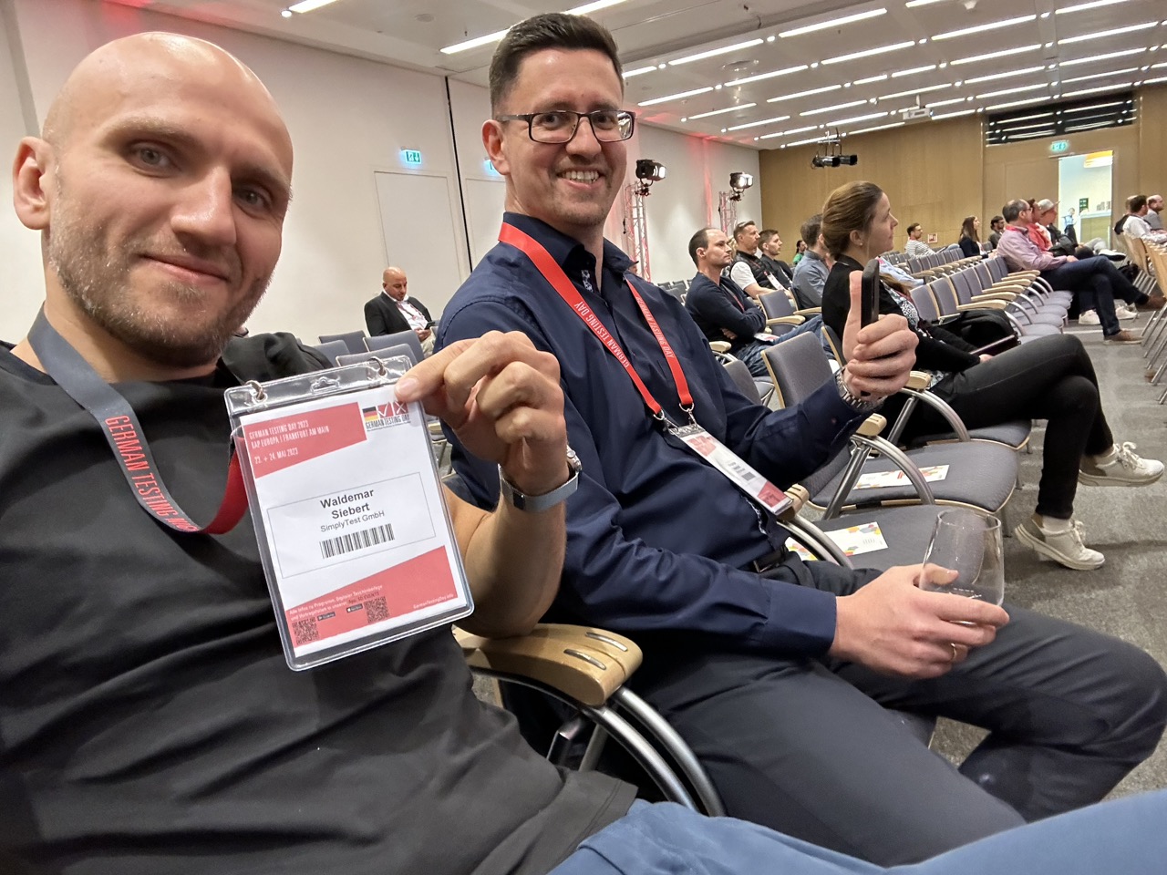 Zwei Kollegen von SimplyTest mit Teilnehmerausweis im Saal des German Testing Day